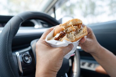 Arabada yemek için hamburger ve patates kızartması tutan Asyalı kadın tehlikeli ve kaza riski taşıyor..