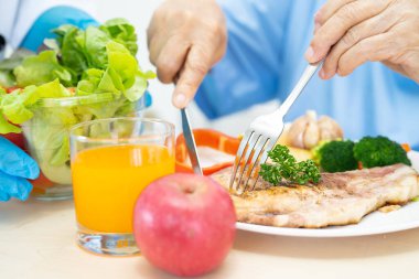 Asyalı yaşlı kadın hasta hastanede yatağında aç ve aç bir şekilde otururken somon bifteği ve sebzeli sağlıklı yemek yiyor..