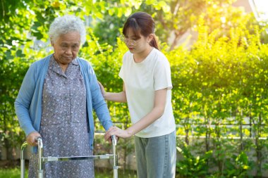 Caregiver help Asian elderly woman patient walk with walker in park, healthy strong medical concept.