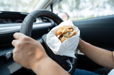Arabada yemek için hamburger ve patates kızartması tutan Asyalı kadın tehlikeli ve kaza riski taşıyor..