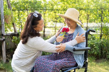 Asyalı yaşlı ya da yaşlı kadın elinde kırmızı gül çiçeği, gülümse ve mutlu bir şekilde güneşli bahçede.