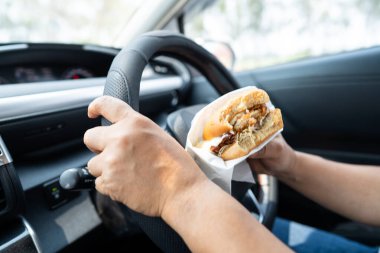 Arabada yemek için hamburger tutan Asyalı kadın tehlikeli ve kaza riski taşıyor..