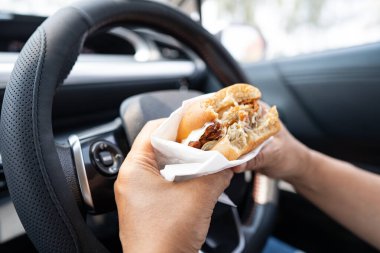 Arabada yemek için hamburger tutan Asyalı kadın tehlikeli ve kaza riski taşıyor..