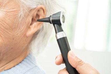 Odiyolog veya ENT doktoru otoskop kullanarak işitme sorunu olan Asyalı yaşlı kadın hastaların kulaklarını kontrol eder..