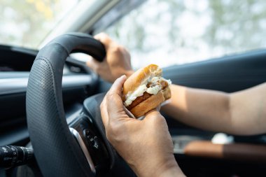 Arabada yemek için hamburger tutan Asyalı kadın tehlikeli ve kaza riski taşıyor..