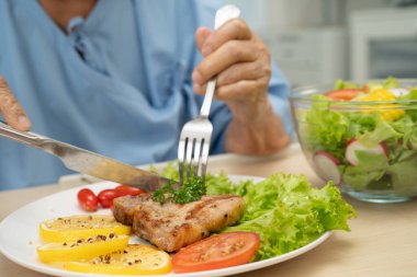 Asyalı son sınıf kadın hasta hastanede sağlıklı yemek için domuz pirzolası ve sebze salatası yiyor..
