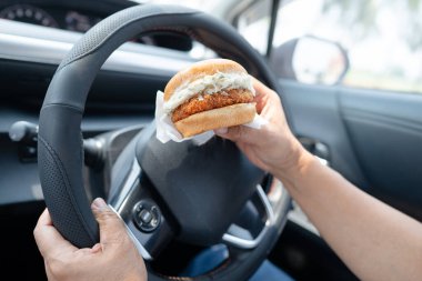 Arabada yemek için hamburger tutan Asyalı kadın tehlikeli ve kaza riski taşıyor..
