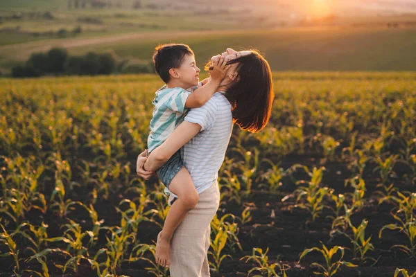 Happy young caucasian mother and her son having fun outdoor on a summer day in a green corn field over sunset. Mother nature. Summer nature. Fun family. Natural