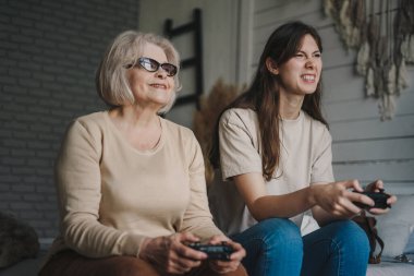 Yaşlı anne, yetişkin kızıyla evde video oyunu oynuyor, kanepede oturup joystick kullanıyor. Mutlu bir aile. Aile içi hobilerinin keyfini çıkarıyorum.