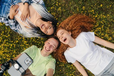 Şehir parkında çimlerin üzerinde uzanan ve cep telefonuyla kendi portresini çeken üç arkadaşın en iyi görüntüsü. Kameraya bakıp gülümse. Seyahat ve aktif yaşam konsepti