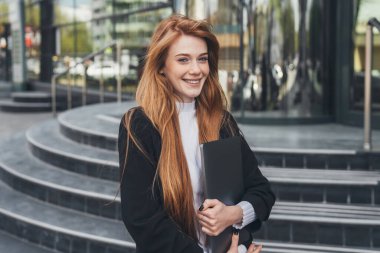 Elinde dizüstü bilgisayarla gülümseyen kızıl saçlı iş kadını yemek molasında ofis binasının önünde kameraya gülümsüyor. Teneffüste dışarıda. İş dünyası