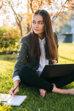 Genç beyaz bir kadın parkta dizüstü bilgisayar kullanıyor, açık havada ders çalışıyor çimlerin üzerinde dizüstü bilgisayarla oturuyor. İnternet teknolojisi. Yaz tatili. Sörf yapmak