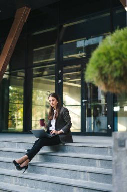 Ofis merdivenlerinde oturmuş kahve fincanı içerken dizüstü bilgisayar kullanan ceketli güzel bir kadının görüntüsü. Teneffüste kahve.