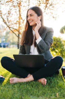 Parktaki yeşil çimlerde oturan, yaz günü bacak bacak bacak üstüne atmış ve dizüstü bilgisayarla çalışan güzel bir kadının portresi. Eğitim internet teknolojisi