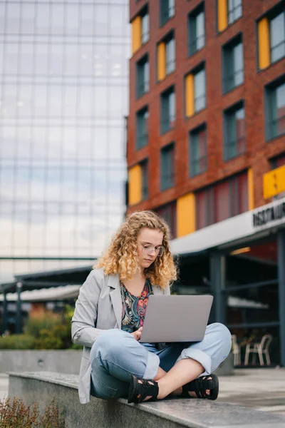 stock image Beautiful and young girl with curly-hair and eyeglasses studying with a laptop, surfing the internet, using multimedia content and devices outside the college