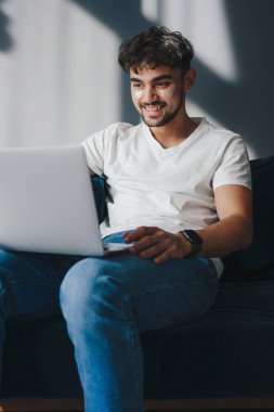 Göz altında yamaları olan meşgul bir adam, kilitlenirken evde dizüstü bilgisayarla çalışıyor, dizüstü bilgisayar ekranına bakarken gülümsüyor. Cilt bakımı güzeli. Gerçek hayat. Günlük
