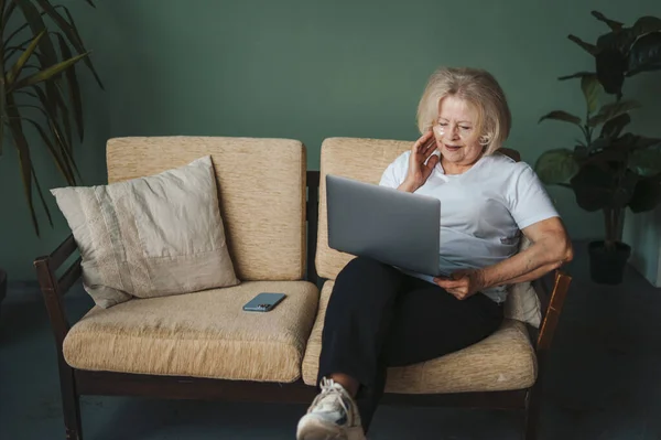 Caucasian senior woman with under eye patches sitting on sofa in front of laptop during remote work at home. Internet technology. Skin care.
