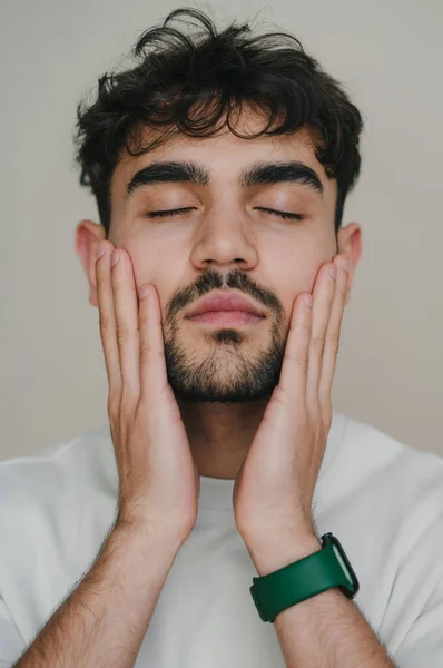 Close-up portrait of a man with eye closed touching his face with hands confidently standing in front of the camera. Feeling calm and confident. Concept of skin
