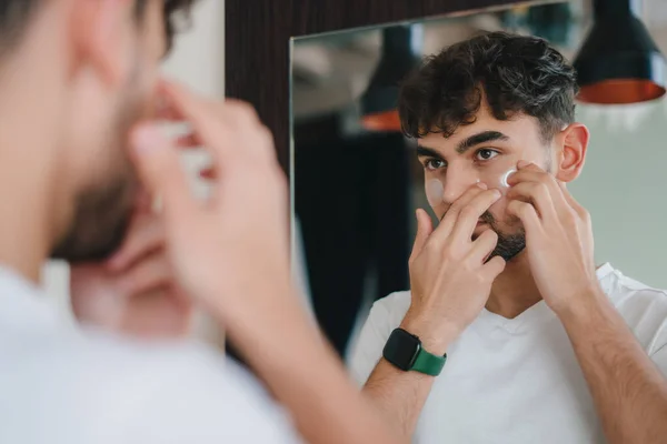 Portrait of a man in a white T-shirt applying patches under eyes caring for face skin looking in mirror. Beauty treatment. Dermatology, cosmetology. Eye care