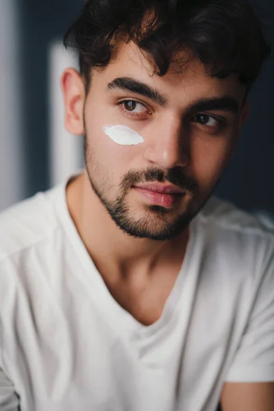 Man portrait of a handsome young man applying moisturising cream to his face, looking away. Beauty portrait. Man portrait. Beauty routine. Skin care routine.
