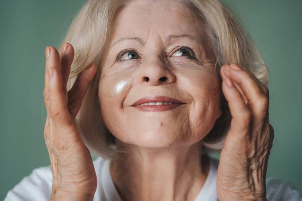 Close-up portrait of a retired woman doing cosmetics procedures with eye patches under eyes isolated over green wall. Dry skin care. Beauty therapy concept
