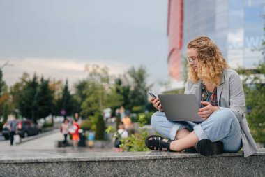 Student woman with curly-hair studying on smart phone and laptop computer at university campus. Free space for text. Distance education. clipart