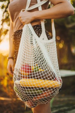 Elinde tekrar kullanılabilir meyve ve sebzelerle bahçede yürüyen bir kadın kolunun görüntüsü. Sıfır atık. Paketsiz yiyecek alışverişi. Eko