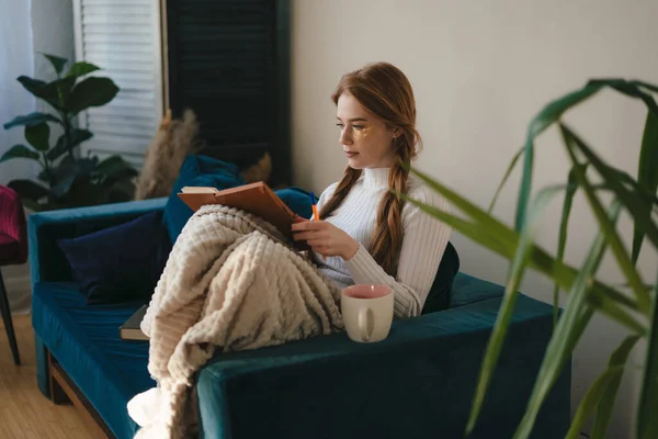 Beautiful woman with cosmetic eye patches sitting on the sofa in cozy clothes and writing in notebook. Morning habits and home beauty routine. Self care.