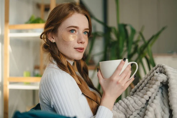 Young beautiful freckled woman with eye patches drinking morning coffee on sofa. Morning habits and home beauty routine. Beauty treatment. Female beauty routine