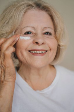 Gülümseyen yaşlı bir kadının portresi. Gözlerinin altındaki siyah halkaları çıkarmak için yaşlanma önleyici losyon sürüyor. 50-60 yıl sonra cilt bakımı. Sabah güzellik rutini.