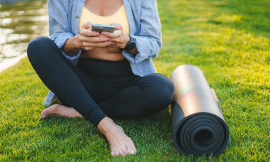 Spor kadın telefonu tutuyor yoga minderinin yanındaki yeşil çimlerin üzerinde oturuyor, sabah antrenmanından sonra dinleniyor. Antrenman egzersizi. Fitness kadını. Sağlıklı yaşam tarzı, spor