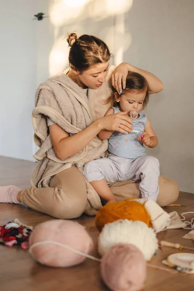 Smiling young Caucasian mother sitting on the floor, knitting with needles with her cute little baby girl daughter. Improve motor skills with small girl child