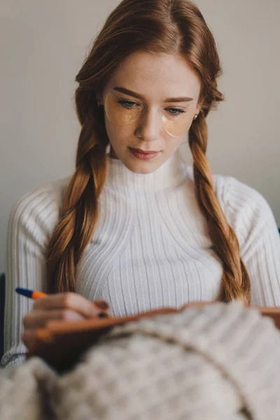 一个红头发女人的画像 辫子插在两条辫子里 坐在沙发上的毛毯下 边写诗边用小记事本记笔记 创意过程 — 图库照片