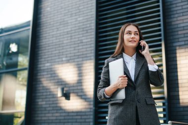 Şehir caddesinde dizüstü bilgisayarı olan ve cep telefonuyla arama yapan çekici bir iş kadınının portresi. Uzayı kopyala Modern yaşam tarzı. Boşluğu kopyala