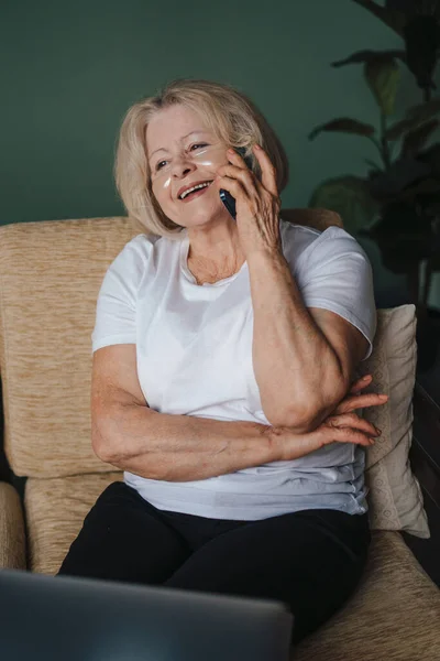 Senior woman with eye-patches talking on phone and using laptop in living room. Family home leisure. Internet communication, wireless connection technology.