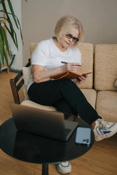 Portrait of elderly focused grey-haired woman sitting on sofa, making notes in notepad at home with open laptop. Internet communication. Education internet