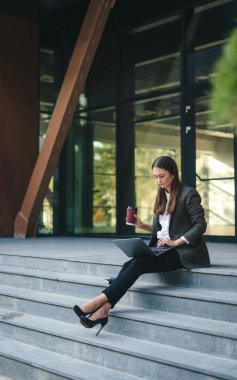 İş kadını dışarıda dizüstü bilgisayar kullanıyor ofis binasının yanında oturuyor ve uzaktan kumanda yapıyor. Serbest çalışan serbest çalışan bir işçi. Veri analizi Yazma bilgisi.