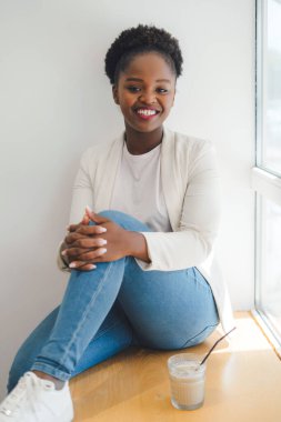 Kafenin pencere eşiğinde oturan neşeli, genç Afrikalı bir kadın. Kahve dükkanında kahve içen bir kadın. Modern teknoloji. Pozitif kişi. Güzellik