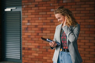 Güzel kıvırcık saçlı bir öğrenci elinde defterlerle dışarıda, şehir sokağında poz veriyor. Üniversite öğrenim programları, akademik eğitim reklamları