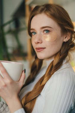 Ginger, güzel, genç bir kadın, kanepede oturuyor ve sabahları bir fincan kahve içiyor. Sabah rutini konsepti. Konforlu daire.