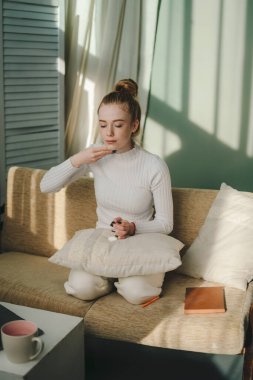 Beyaz tenli güzel bir kadın evde otururken yüzüne krem sürüyor. Cilt bakımını düşün. Samimi bir daire. Dairenin içi. Cilt bakımı. Vücut bakımı.