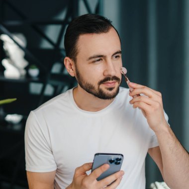 Akıllı telefonu elinde tutarken ve yanağına yeşim taşlı bir masaj aleti takarken yüzünü çeviren sakin bir adamın portresi. Dermatoloji, kozmetoloji. Vücut bakımı