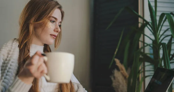 Happy young ginger woman with eye-patches under eyes drinking coffee covered with blanket while sitting on sofa at home watching movie using laptop. A cozy