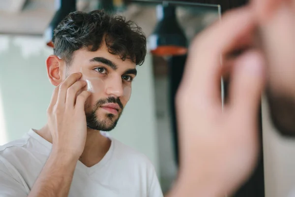 Bearded man applying eye patches under his eyes while looking at mirror. Wrinkles and face home care for men. Skin care routine. Home spa care concept. Facial