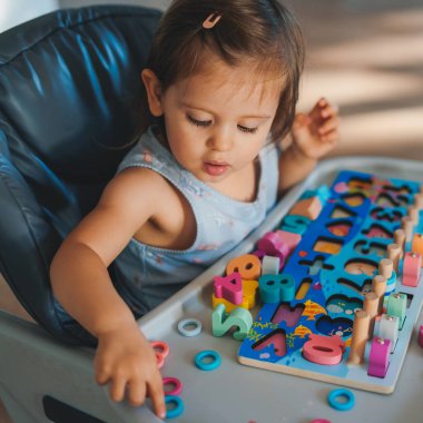 Renkli gökkuşağı oyuncağıyla oynayan sevimli bir kız bebek. Sandalyede oturuyor. Harflerle ve sayılarla gelişen bir ahşap oyun.