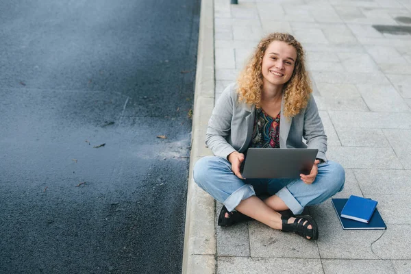 Σγουρομάλλα Φοιτήτρια Κάθεται Στο Δρόμο Βάζοντας Laptop Στα Πόδια Και — Φωτογραφία Αρχείου