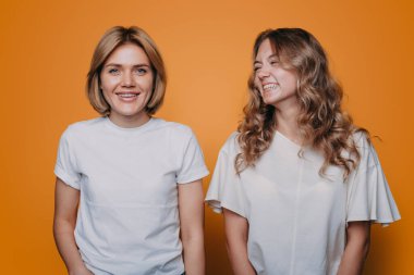 Pozitif iki genç kadın, turuncu arka planda gelişigüzel giyinip birbirlerine yakın durarak eğleniyorlar. İnsan yaşam tarzı.