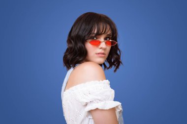 Ön manzara portresi. Kırmızı güneş gözlüklü güzel bir kadın. Kameraya bakıyor. Beyaz yaz elbiseli çekici bir kadın, maviliğe karşı duruyor.