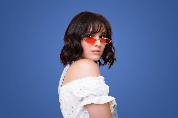 Портрет с видом спереди красивая молодая женщина в красных солнцезащитных очках, смотрящая через плечо на камеру. Привлекательная женщина в белой летней одежде, стоящая против синей