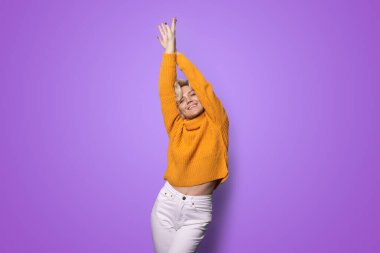 Güzel, tatlı, rahat, çekici bir kadın mor arka planda poz veriyor, kollarını kaldırıyor, kameraya bakıyor..